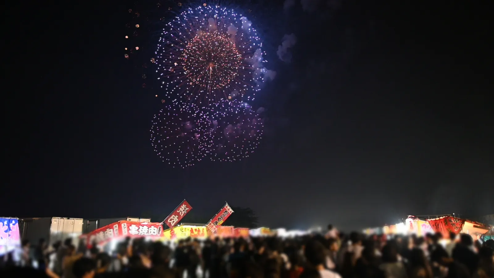 ふくしま花火大会で露店に並ぶ人混みの上に花火が見える様子